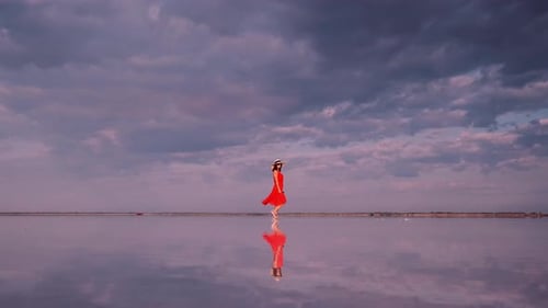 Young Woman in a Waving Dress Walks Along a Pink Lake in Which Clouds Are Reflected.
