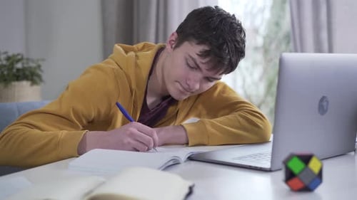 Teenager Writes in Notebook Near Laptop at Desk