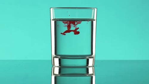 Drop of Red Ink Dripping Into Clear Water in Glass Standing on Turquoise Background in Slowmotion