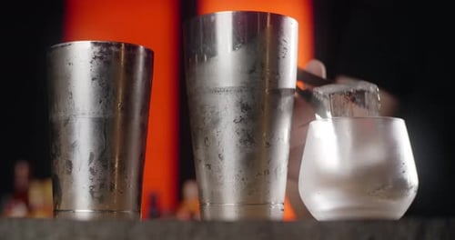 Bartender Puts Big Ice Cube to the Frozed Cocktail Glass By Forceps Making the Cocktail in the Bar