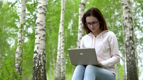 Young Woman in Nature with a Laptop in Her Hands. A Girl Sits on a Stump in a Birch Forest and Leads