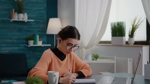 Woman Writing in Notebook at Desk with Laptop