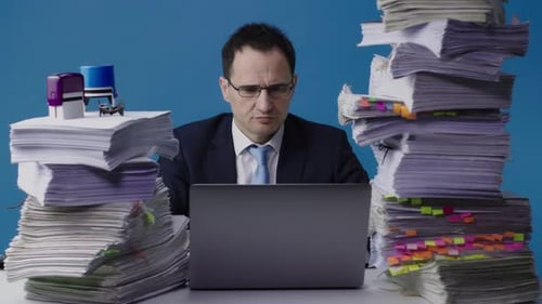 Serious Manager Working on Laptop Among Piles of Paperwork, Overloaded with Work