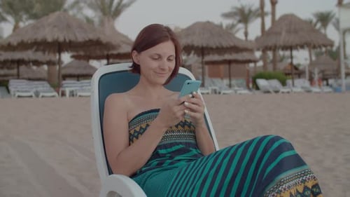 Woman Typing Message Holding Cell Phone in Hands Sitting on the Beach Chair By the Sea