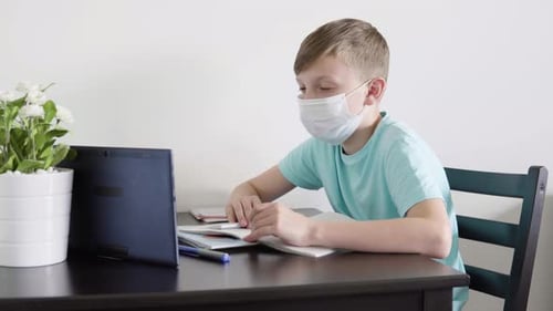 A Young Boy in a Face Mask Talks To Someone on a Tablet As He Does Homework for School at a Table