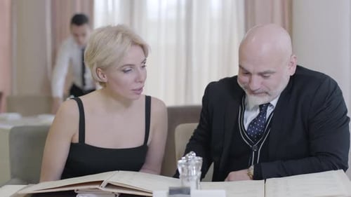 Positive Caucasian Man and Woman Discussing Order in Restaurant with Blurred Waiter Cleaning Table