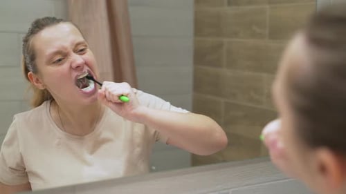 Woman in Beige Tshirt Actively Brushes Her Teeth with Green Toothbrush in the Bathroom at Home