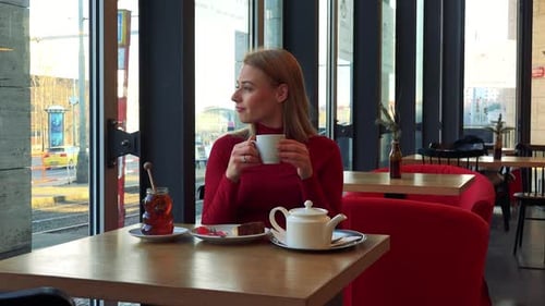 A Young Attractive Woman Sits at a Table with Meal in a Cafe, Drinks Tea and Looks Out a Window