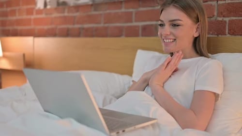 Fröhliche junge Frau macht Video-Chat auf Laptop im Bett