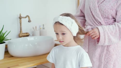 Mother brushing little girl's hair in bright bathroom