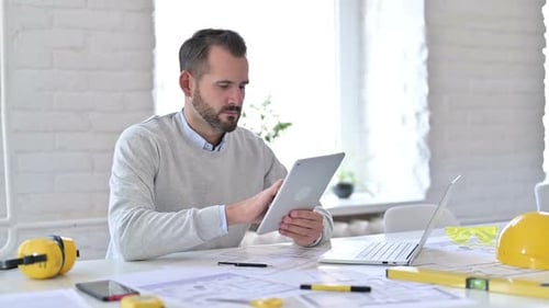 Junger Architekt mit Laptop nutzt Tablet im Büro