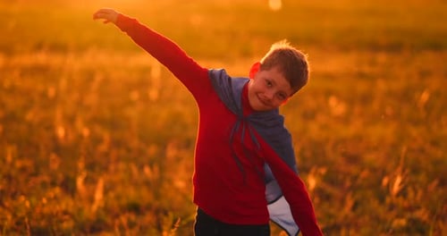 A Child in the Costume of a Superhero in a Red Cloak Runs Across the Green Lawn Against the Backdrop