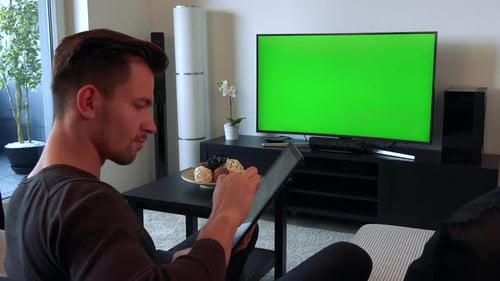 A Young, Handsome Man Watches a TV with a Green Screen in a Cozy Living Room and Types on His Tablet