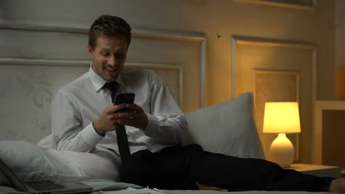Smiling Man Is Suit Sitting on Bed and Texting on Smartphone, Dating Site, App