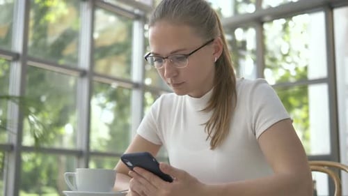 Woman with Glasses Surfs Internet with Mobile Phone in Cafe