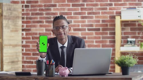 Black Man Office Worker in Glasses Smiles Shows Green Phone Screen with an App and Sits at a Desk