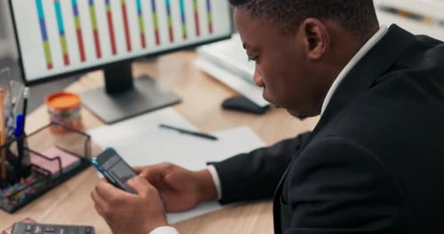 A Darkskinned Man Dressed in a Suit Sits at a Desk in Front of a Computer Monitor Holding a Phone in