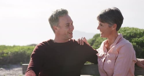 Caucasian couple enjoying free time by sea on sunny day sitting on bench and talking