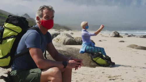 Senior hiker man wearing face mask with backpacks sitting on a rock and senior woman taking pictures