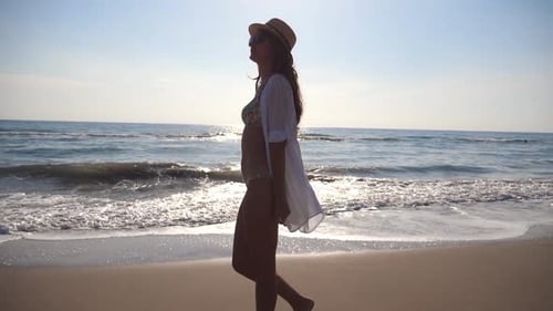 Happy Woman in Bikini and Shirt Walking on the Beach Near the Ocean. Young Beautiful Girl in Hat