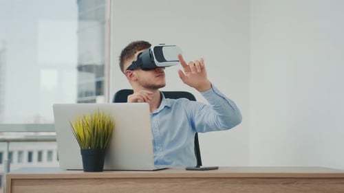 A Young Man Sitting at a Desk in the Office Uses Augmented Reality Glasses To Work on Business