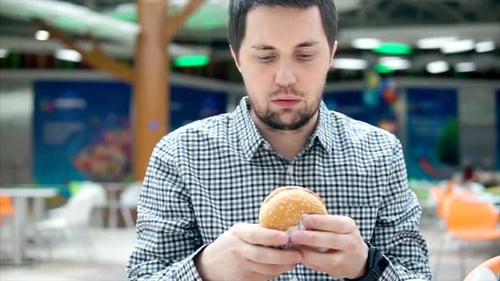 A Young Man Is Eating a Hamburger with Meat in a Food Court.