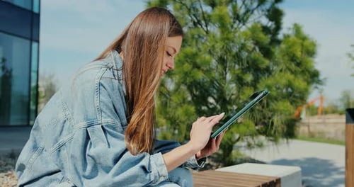 Woman is Sitting on a Park Bench and Scrolling on the Tablet in Her Hands