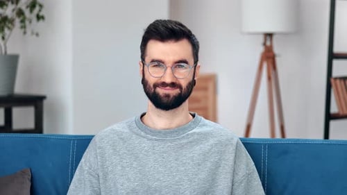 Nahaufnahme Porträt Brutaler bärtiger Mann in Brille trägt graues trendiges Sweatshirt posiert auf der Couch