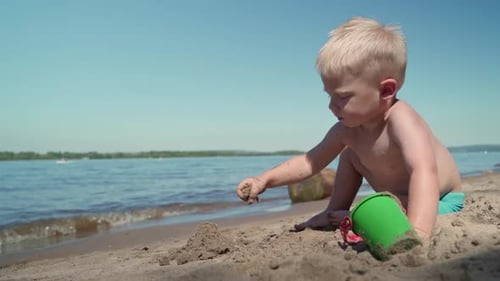A little boy sits on the shore of the lake and plays in the sandbox.