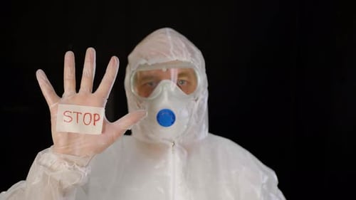 Medizintechniker in Schutzanzug, Maske und Handschuhen zeigt Stop-Corona-Virus auf schwarzem Hintergrund