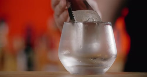 Bartender Puts Big Ice Cube to the Frozed Cocktail Glass By Forceps Making the Cocktail in the Bar