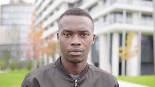A Young Black Man Looks Seriously at the Camera - Closeup - Office Buildings in the Background