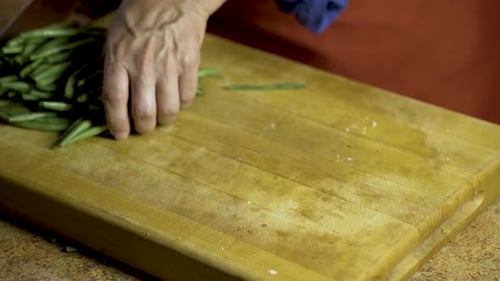 Koch, reiht einen Stapel frischer grüner Bohnen auf und schneidet sie auf einem Holzschneidebrett in zwei Hälften.
Kamera