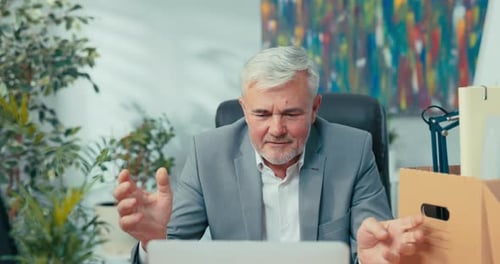 Man in Suit with Gray Hair Leads Remote Conversation From Office Sitting at Desk in Front of Laptop