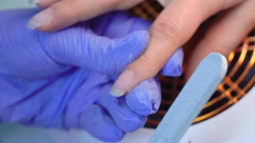 Close-up of manicure master in blue gloves filing nails with special nail file.