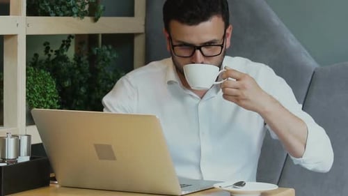 Handsome Businessman Working on His Computer and Drinking From White Cup in Cafe
