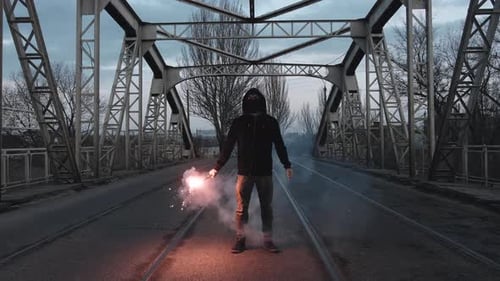 Young Man in Hoodie and Balaclava with Red Burning Signal Flare on the Road Under an Old Steel Frame