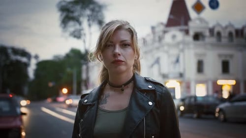 Tattooed Young Woman Hipster in Black Leather Jacket and Choker is Standing Near a Road of the City
