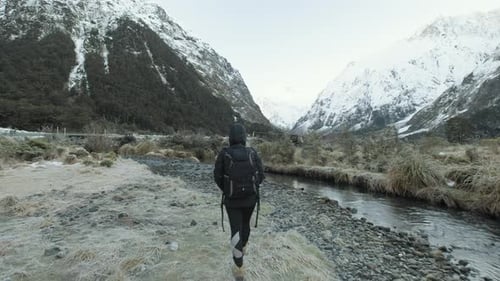 Girl in hiking gear walking along river surrounded by snow capped mountains