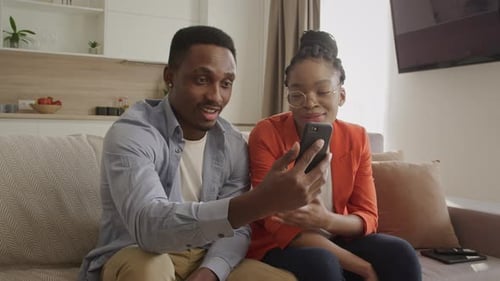 Young Adult African American Couple Sitting on a Sofa and Waving Their Friends During a Video Call
