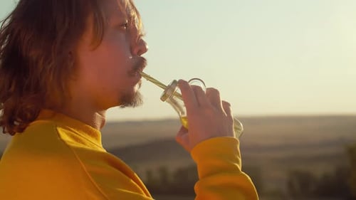 Happy Bearded Man with Long Hair Drinks Cocktail Against Background of Field and Smiling Dances