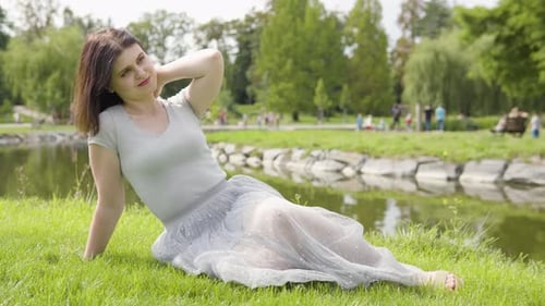 A Young Caucasian Woman Enjoys the Sun with a Smile As She Sits in Grass By a Pond in a City Park