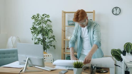 Young Architect Looking at Blueprint and Then at Computer Screen Working at Home Alone
