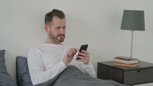 Man Lying in Bed Looking Upset at Phone