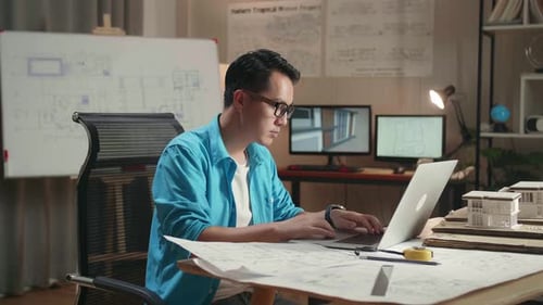 Asian Man Engineer Looking At Blueprint And Typing On A Laptop Before Walking Out Of The Office