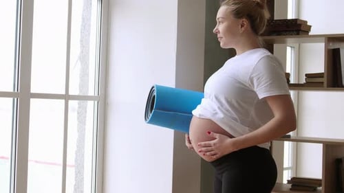 Pregnant Woman Holding Yoga Mat Indoors by Window