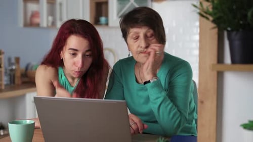 Woman Helps Senior Woman Use Laptop in Kitchen