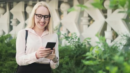 Beautiful business woman in a shirt, black skirt and glasses in a park with green bushes holds a mob