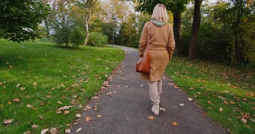 A Woman in a Coat with a Bag Walks in an Autumn Park Rear View