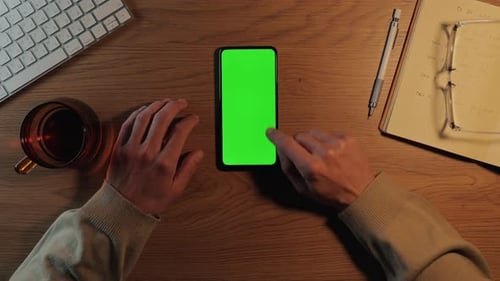 Homem tocando na tela verde do celular enquanto está sentado na mesa